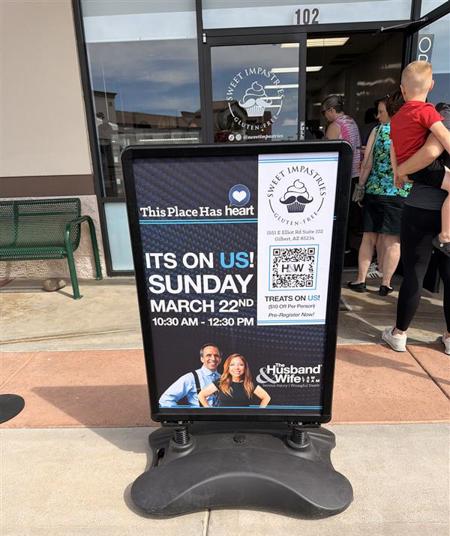 A Husband & Wife Law Team sign stands outside Sweet Impastries, promoting a free March 22nd event with treats and a QR code, as customers line up at the bakery.