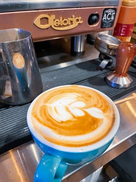 A blue cup of latte with latte art on a barista counter, surrounded by a milk frother and a tamper.