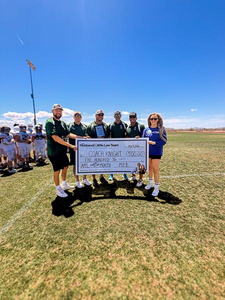 Football coaches gather on the field holding a $500 ceremonial check, with young players practicing behind them