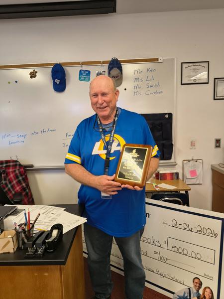 A smiling Mr. Brooks in his classroom, with a large check and awards on the desk behind him.