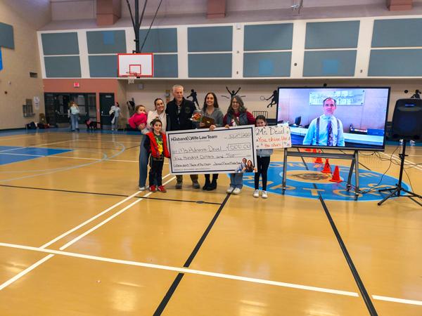 A group of people in a gym holding a large check, standing near a TV screen displaying a person's image. Basketball hoop in the background.