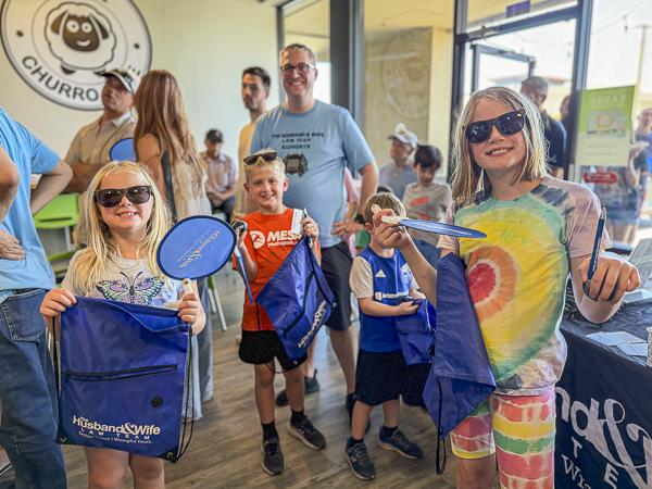 Children in sunglasses hold blue swag bags and fans at the Husband & Wife Law Team’s HEART event, with people and a churro shop logo in the background.
