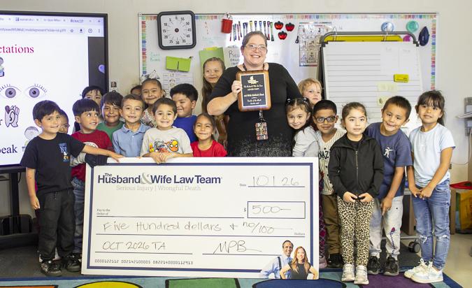 Mrs. Warren celebrates her Teacher of the Month recognition alongside her students, proudly displaying a $500 award and plaque from the Husband & Wife Law Team.