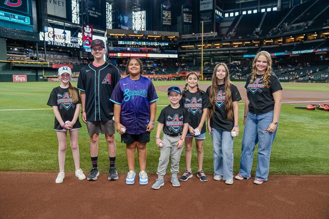 A group of seven people, including children and teens, stand on a baseball field, smiling and holding baseballs, with stadium seating in the background.