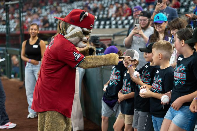Excited children holding baseballs line up for high‑fives from the Diamondbacks mascot, with fans in the Chase Field stands cheering and capturing the moment.