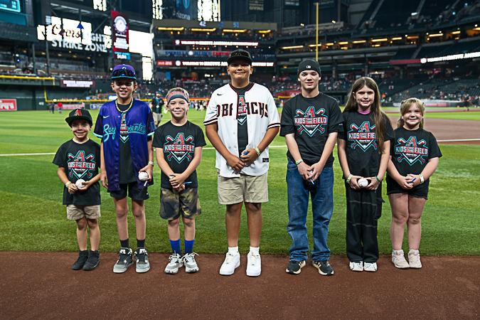 Seven children stand on a baseball field, each holding a baseball. They're smiling and dressed in casual sportswear.