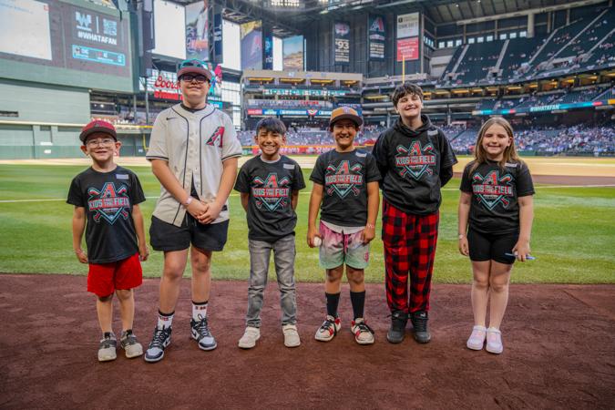 A group of excited kids stand on the field at Chase Field during the Arizona Diamondbacks' 'Kids Take the Field' event. Dressed in team gear, they smile and soak in the moment, with the stadium lights and towering stands surrounding them.
