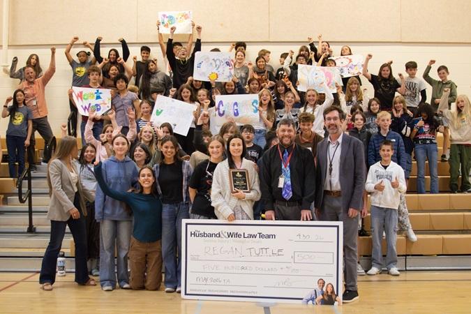 In the gymnasium, students and teachers gather with signs and a large check, with Ms. Tuttle prominently positioned at the front holding a plaque.