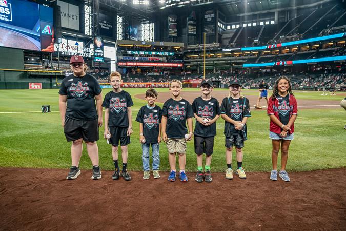 A group of seven children standing on a baseball field, wearing branded shirts and smiling, with the stadium in the background.