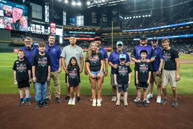 A group of Kids Take The Field winners, including children and adults, stand on Chase Field with stadium lights and the scoreboard glowing behind them.