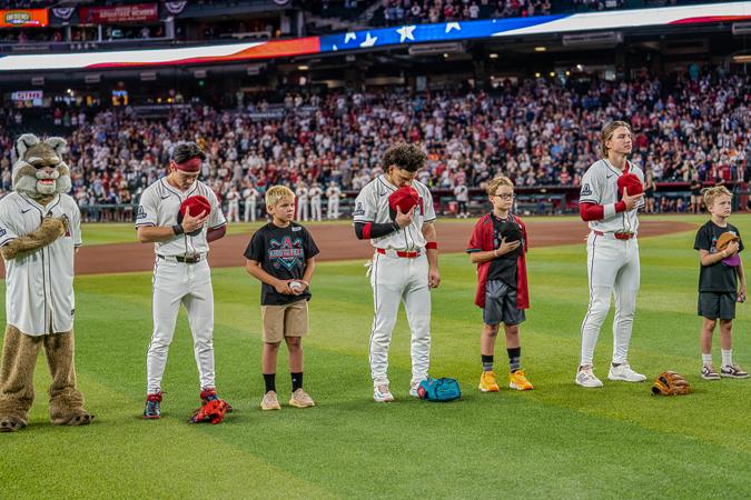 Baseball players and children stand on the field, holding caps over their hearts, during a national anthem ceremony. A mascot stands nearby.
