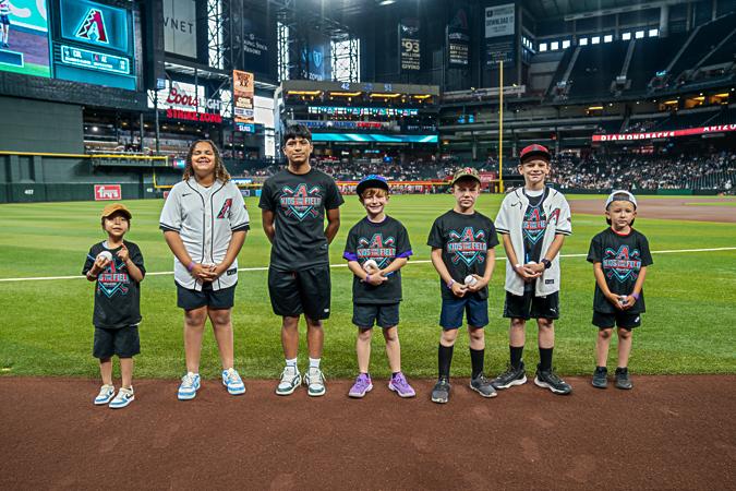 Seven children stand on a baseball field, smiling, wearing sports-themed shirts and hats. Stadium lights and scoreboard visible in the background.