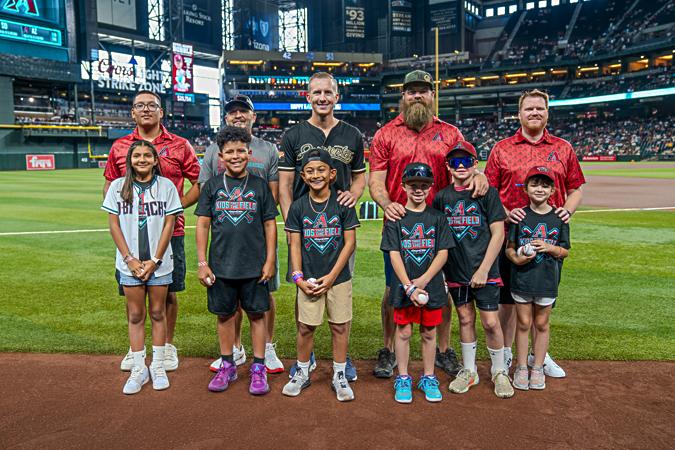 A group of people, including children in baseball attire, pose on a baseball field with a stadium audience in the background.