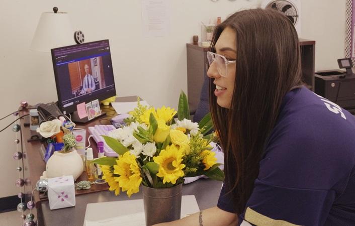 Ms. Branch leans over a student desk in her classroom, yellow and white flowers beside her, as Mark Breyer congratulates her during a Zoom call.